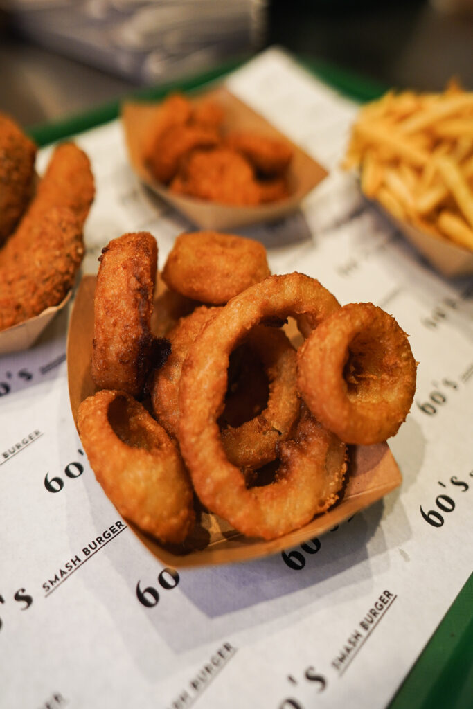 Knusprige Zwiebelringe, Hähnchennuggets und Pommes frites, serviert in Pappschalen auf 60's Smash Burger Papier