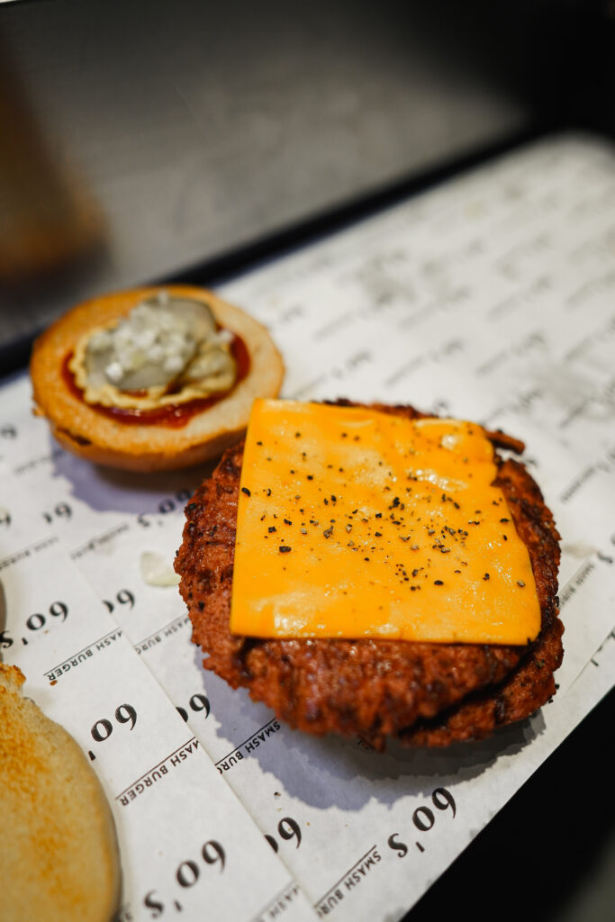 Ein Burger-Patty mit geschmolzenem Cheddar-Käse und schwarzem Pfeffer, daneben eine Brötchenhälfte mit Sauce, Gurkenscheiben und Zwiebeln