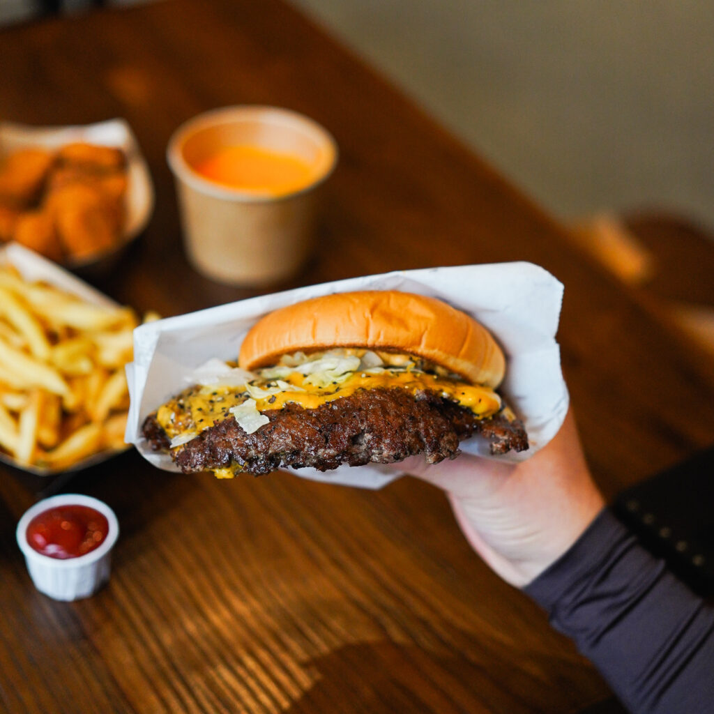 Ein saftiger Cheeseburger mit geschmolzenem Käse und Zwiebeln, gehalten in der Hand, im Hintergrund Pommes, Nuggets und Käsesauce