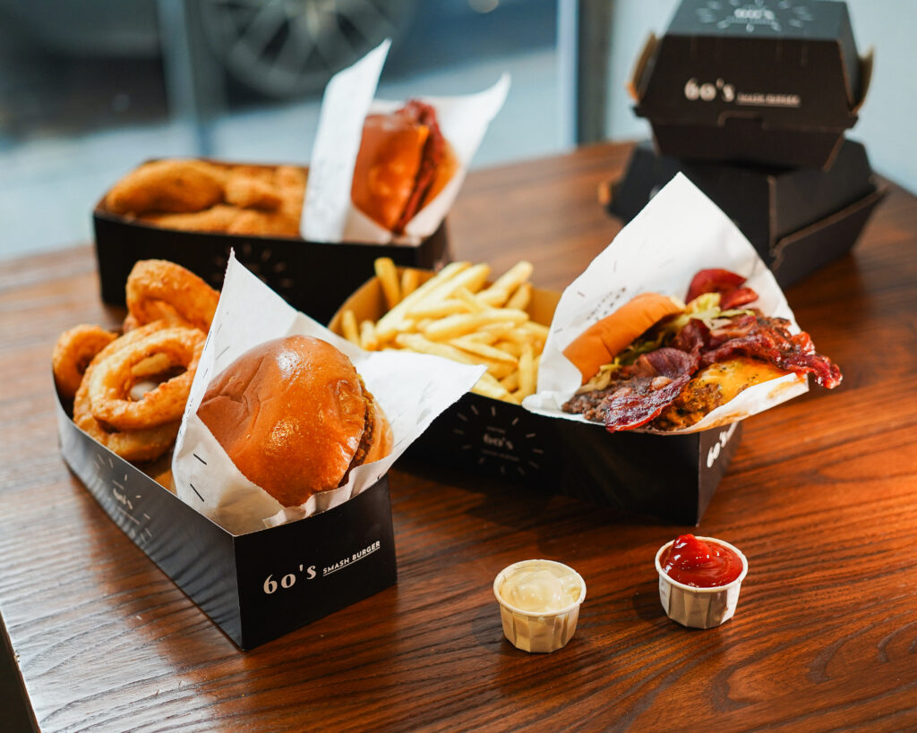 Vielfältige Menüauswahl mit Burgern, Pommes, Onion Rings und Saucen, serviert in 60's Smash Burger Boxen